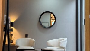 Habitació moderna amb dues butaques blanques, una taula rodona i un mirall rodó a la paret.