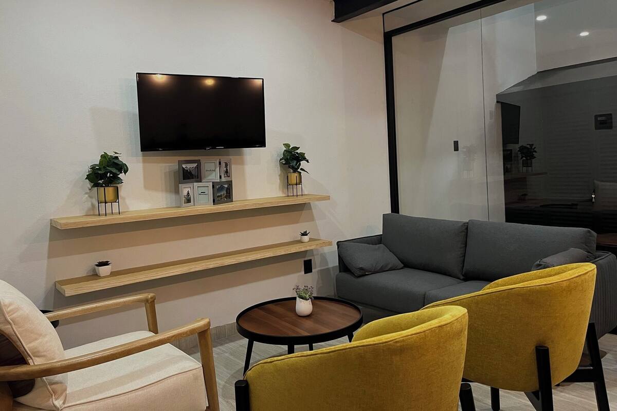 A modern living room with a flat-screen TV mounted on a wall, a grey sofa, yellow armchairs, a wooden coffee table, and a small potted plant.