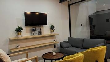 A modern living room with a flat-screen TV mounted on a wall, a grey sofa, yellow armchairs, a wooden coffee table, and a small potted plant.