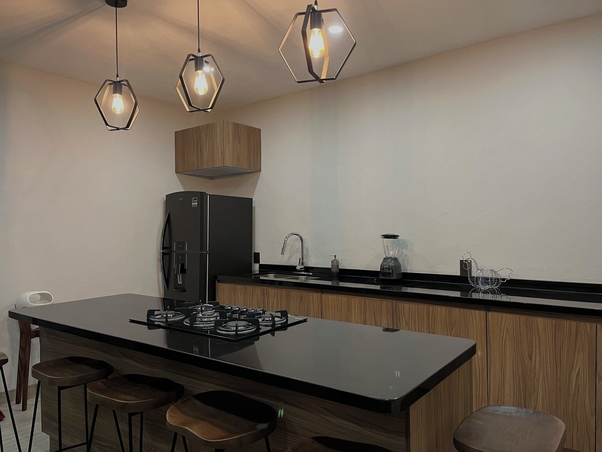 Una cocina moderna con una isla central, taburetes de bar, una estufa a gas empotrada, un refrigerador negro y lámparas colgantes hexagonales.