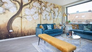 A modern living room with a large wall mural, a blue sofa set, a glass coffee table, and a mustard-colored bench. - Palm Tree House | Birds of Feather (Greater London)