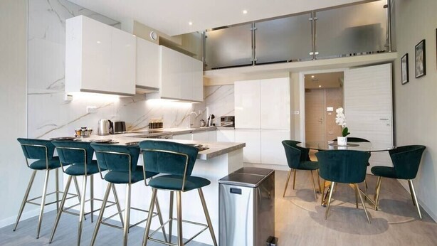 A modern kitchen with white cabinets, a central island with bar stools, and a small refrigerator. - Palm Tree House | Birds of Feather (Greater London)