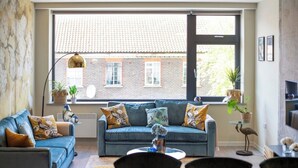 A cozy living room with a blue sofa, a dining table set for two, and a large window overlooking a brick building. - Palm Tree House | Birds of Feather (Greater London)