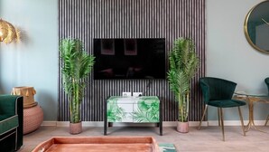 A modern living room with a flat-screen TV mounted on a striped wall, a green sofa, a green armchair, a small coffee table, and two potted plants. - Palm Tree House | Beverly Hills Hotel (Greater London)