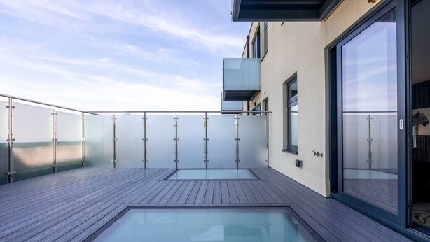 A modern outdoor pool area with a wooden deck and glass walls, providing a view of the sky and surrounding buildings. - Palm Tree House | Panther (Greater London)