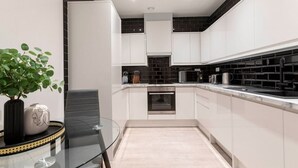 Een moderne keuken met witte kastjes, een zwart betegelde spatwand, roestvrijstalen apparatuur en een glazen tafel met een plant.