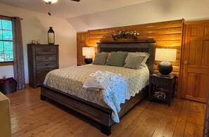 A bedroom with a wooden bed, two bedside lamps, a nightstand, a dresser, and a window with curtains.