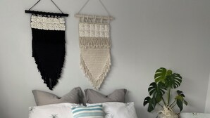 A neatly made bed with a white and blue patterned duvet, a teal throw blanket, and pillows with palm tree prints. There are macramé wall hangings above the bed, a potted plant on a side table, and a floral-patterned chair. - Sun Kissed Studio  (Motueka)