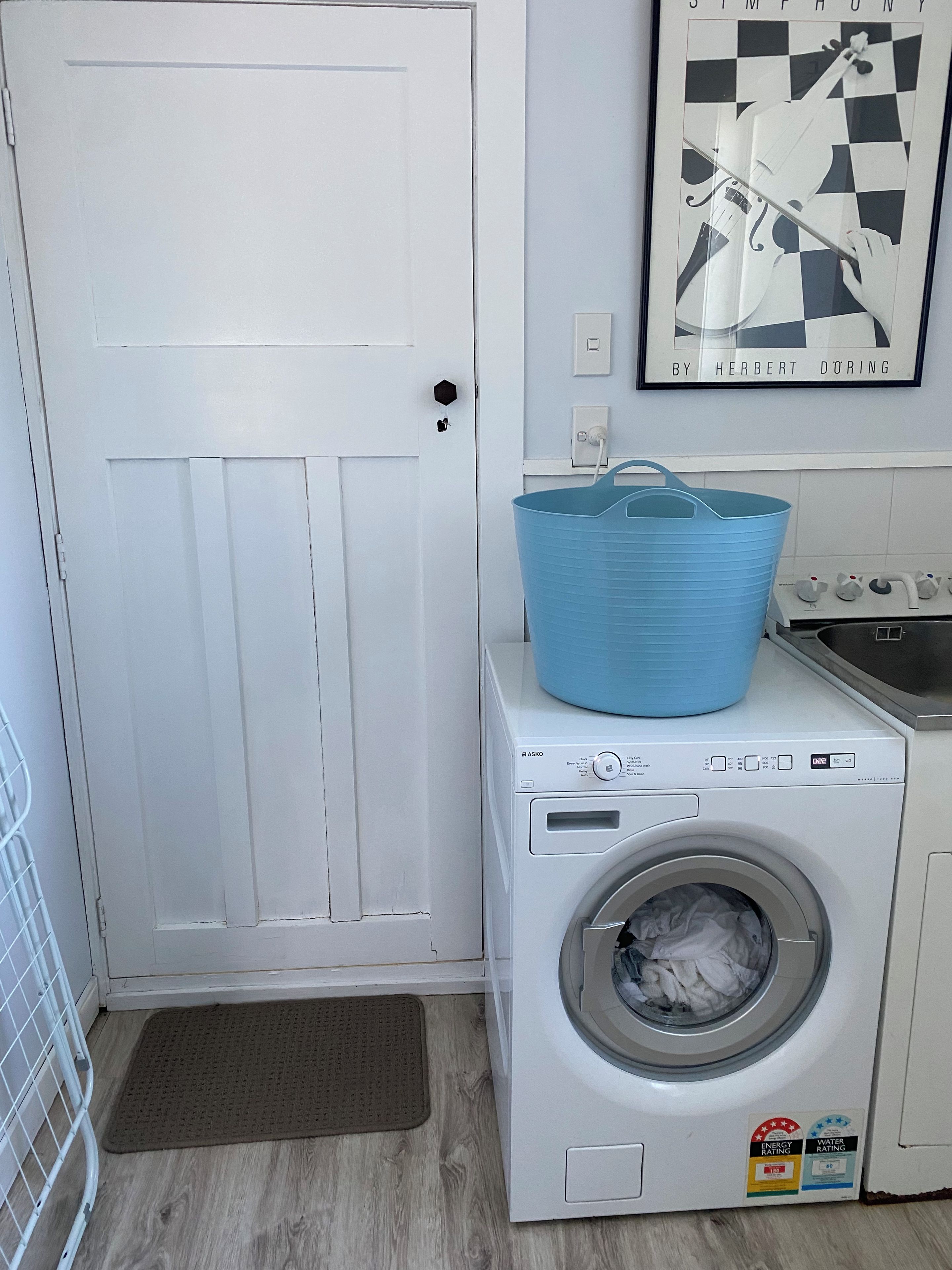 A laundry room with a front-loading washing machine, a blue laundry basket on top, a door, and a framed picture on the wall.