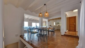 A spacious dining area with a high ceiling and exposed beams, a kitchen in the background, and a wooden door leading to another room. - El Faro - By Almarina Villas (JÁVEA)