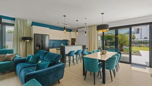 A modern living room with a sofa, dining table, and chairs. There is a kitchen area with a refrigerator and a bar. The room has large windows and a view of the outdoors. - 78 (Medulin)