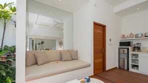 A modern living room with a beige sofa, a wooden door, a kitchen area with appliances, and a large mirror reflecting the room. - Villa Lior (Tabanan)