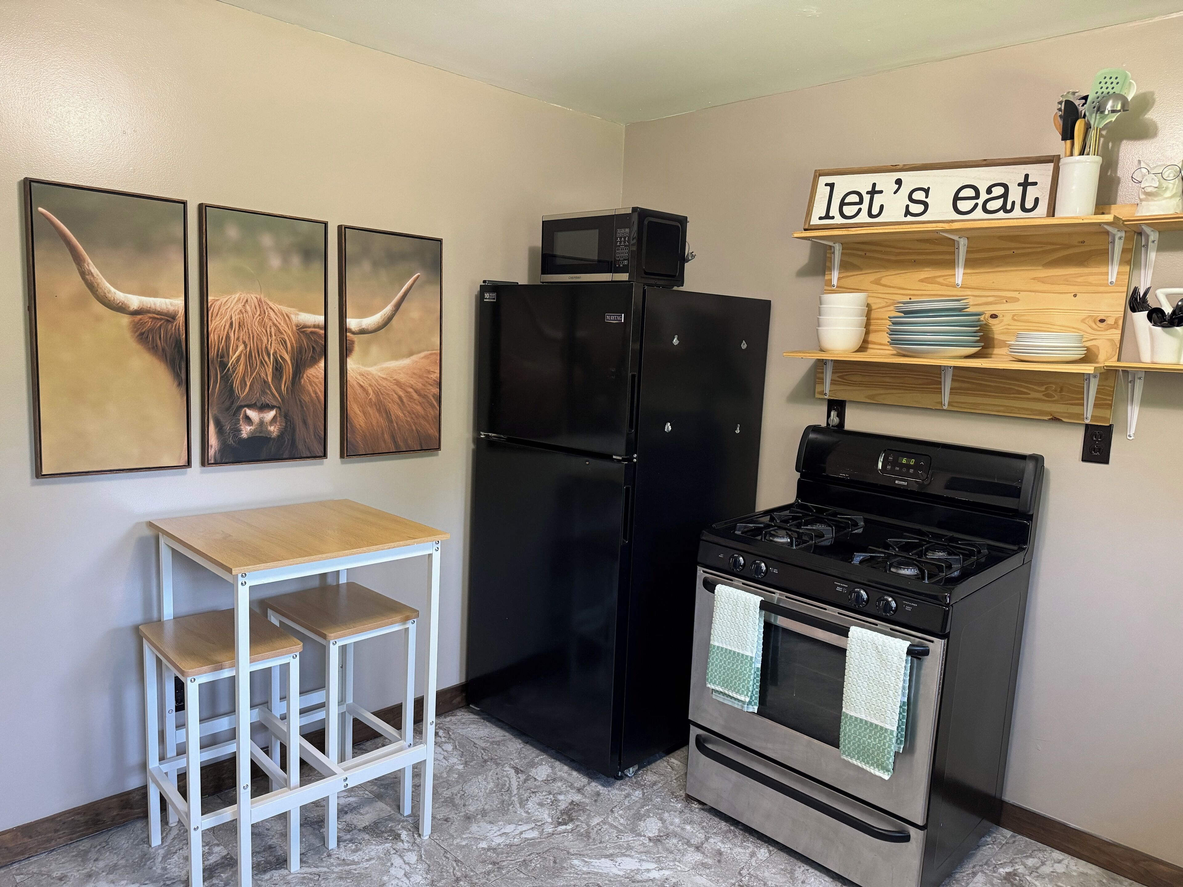 A kitchen with a black refrigerator, a stove, a microwave, and a 'let's eat' sign on the wall. There are three framed pictures on the wall and a small wooden table with two stools.