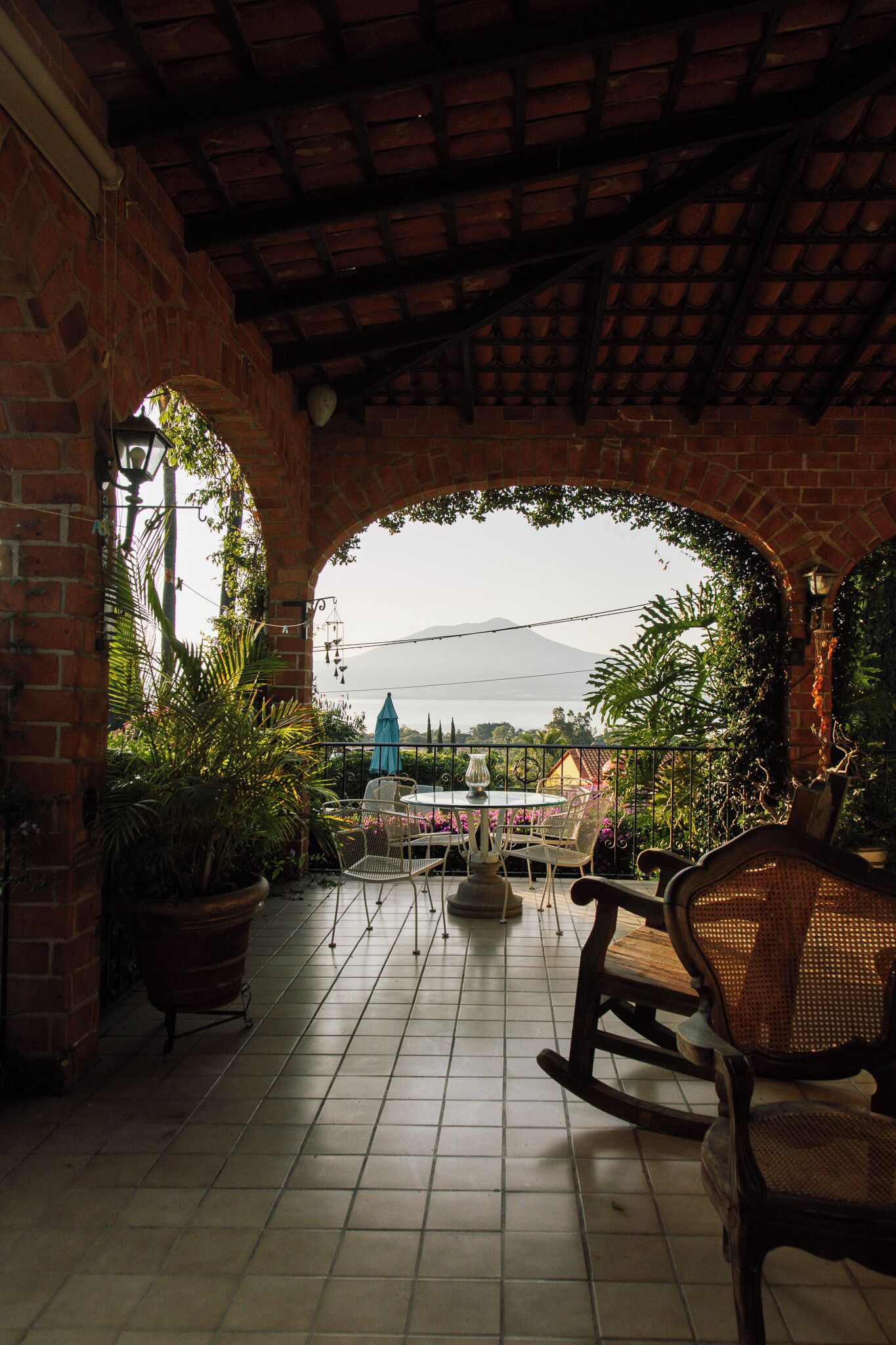 Eine überdachte Terrasse mit Ziegelwänden, Fliesenboden und Bergblick. Es befinden sich Kübelpflanzen, ein Tisch mit Stühlen und ein Schaukelstuhl dort.