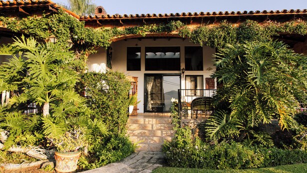 Una casa con tetto in tegole, un vialetto in pietra che conduce all'ingresso e vegetazione rigogliosa, tra cui felci e piante in vaso.