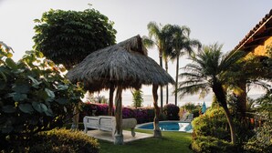 Un gazebo con tetto di paglia, una piscina e vegetazione lussureggiante.