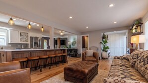A spacious living area with a sofa, ottoman, and bar stools. A kitchen with white cabinets and a refrigerator is visible in the background.