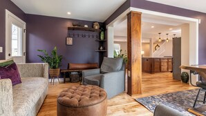 A living room with a sofa, ottoman, and chair, a wooden dining table, and a kitchen in the background.