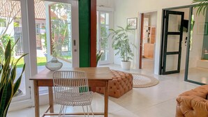 A modern living room with a wooden table, white chair, and a brown sofa. There is a potted plant and a large window with a view of the outside. - LANE HOUSE 1 (Canggu)