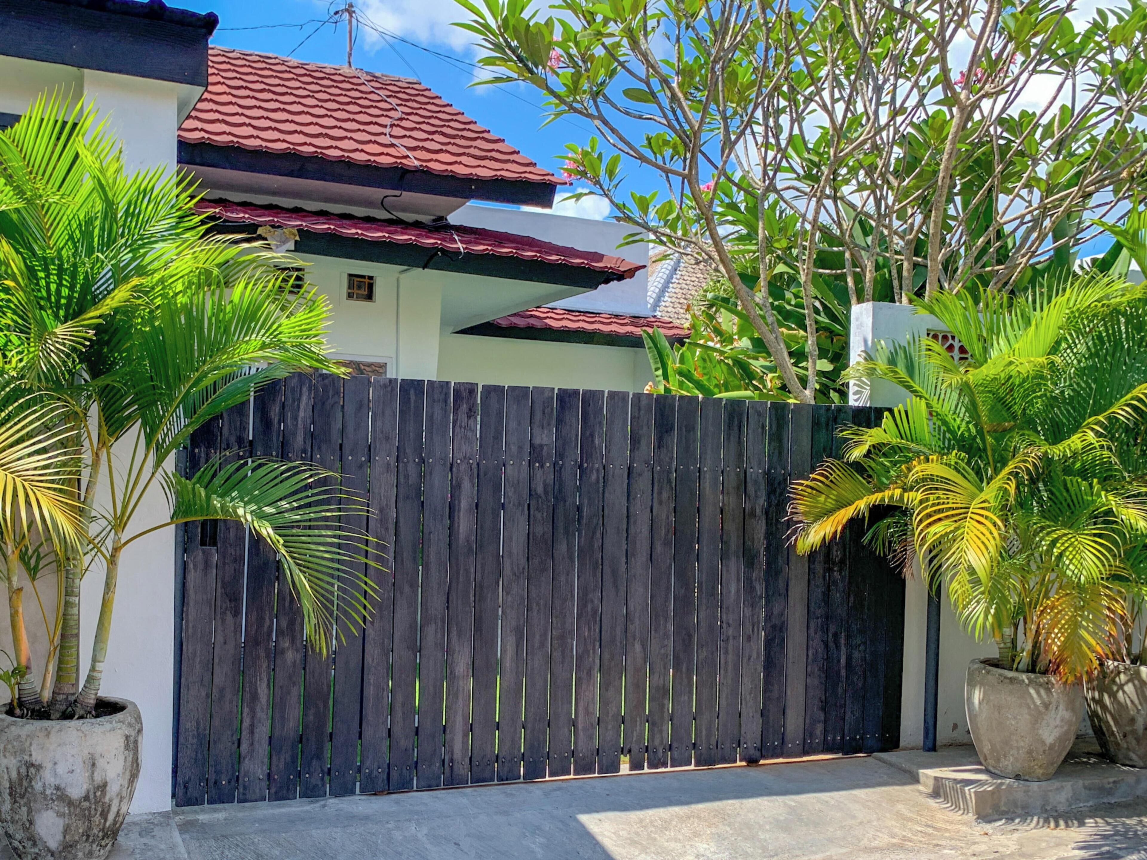 A wooden gate with a concrete pathway leading to a house with a red-tiled roof.
