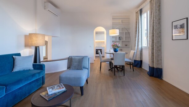 Una sala moderna con un sofá azul, un sillón pequeño y una mesa de centro. Se observa un comedor con mesa y sillas, y una cocina al fondo.