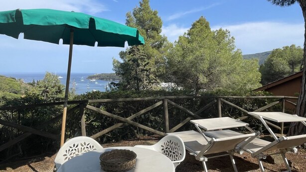 A patio with a green umbrella, white chairs, and a table overlooking a scenic view of the sea and hills. - Appartamento Collina sul Golfo 14 (Porto Azzurro)
