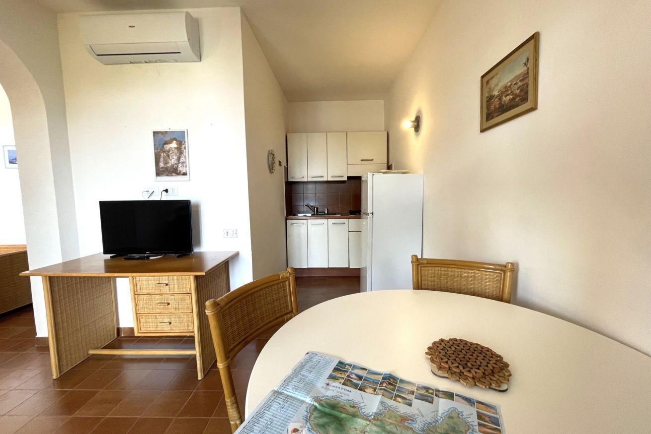 Un espacio habitable funcional con una mesa de comedor, un televisor sobre un escritorio, una zona de cocina y un cuadro en la pared.