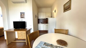 A compact living space with a dining table, a television on a desk, a kitchen area, and a framed picture on the wall. - Appartamento Collina sul Golfo 14 (Porto Azzurro)