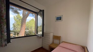 A bedroom with a bed, a window with a view of trees, a dresser, and a chair. - Appartamento Collina sul Golfo 14 (Porto Azzurro)