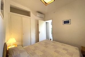 A bedroom with a bed, a nightstand, a lamp, a door, and an air conditioning unit.