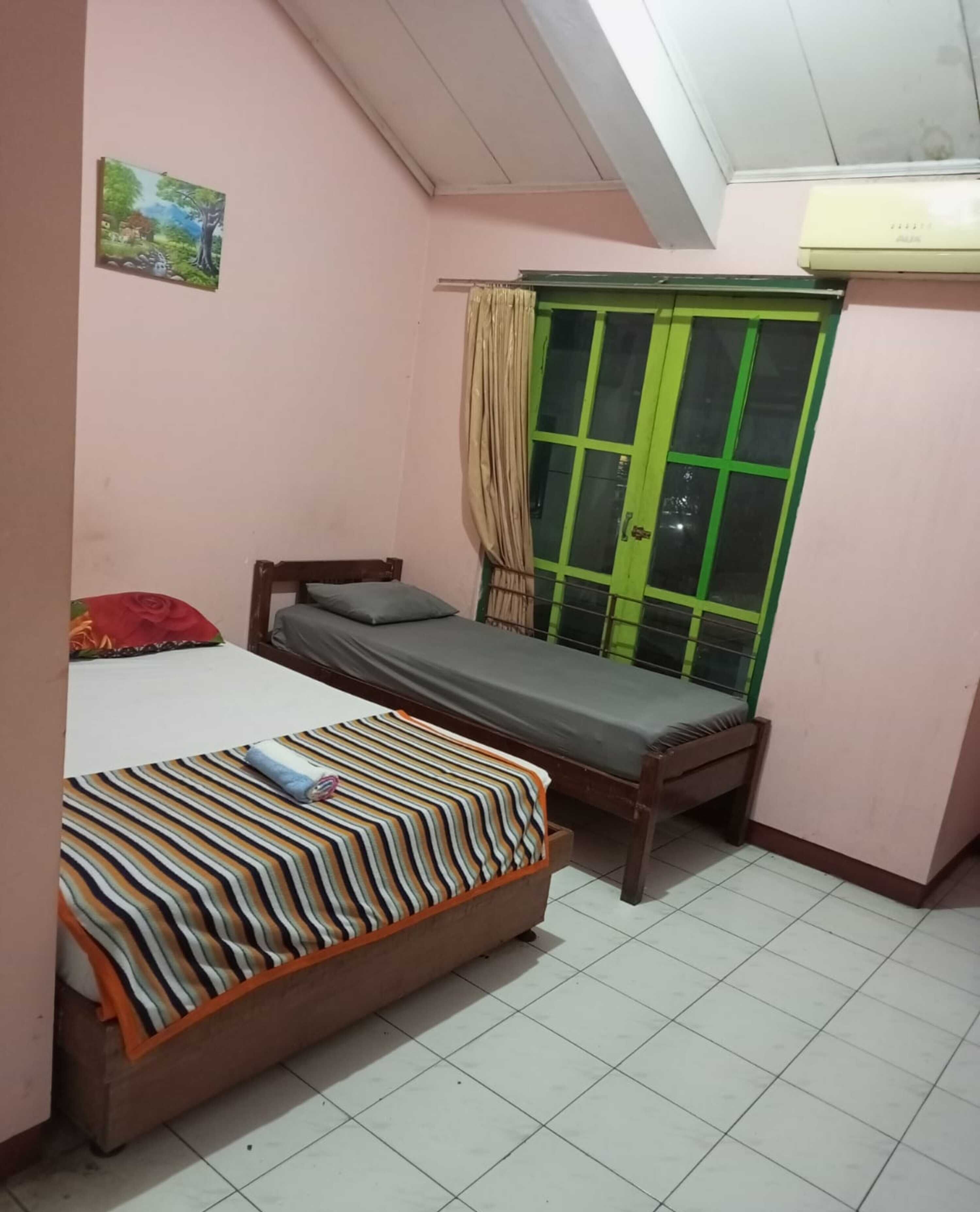 A bedroom with two beds, a green-framed window, a wall-mounted air conditioner, and a small painting.