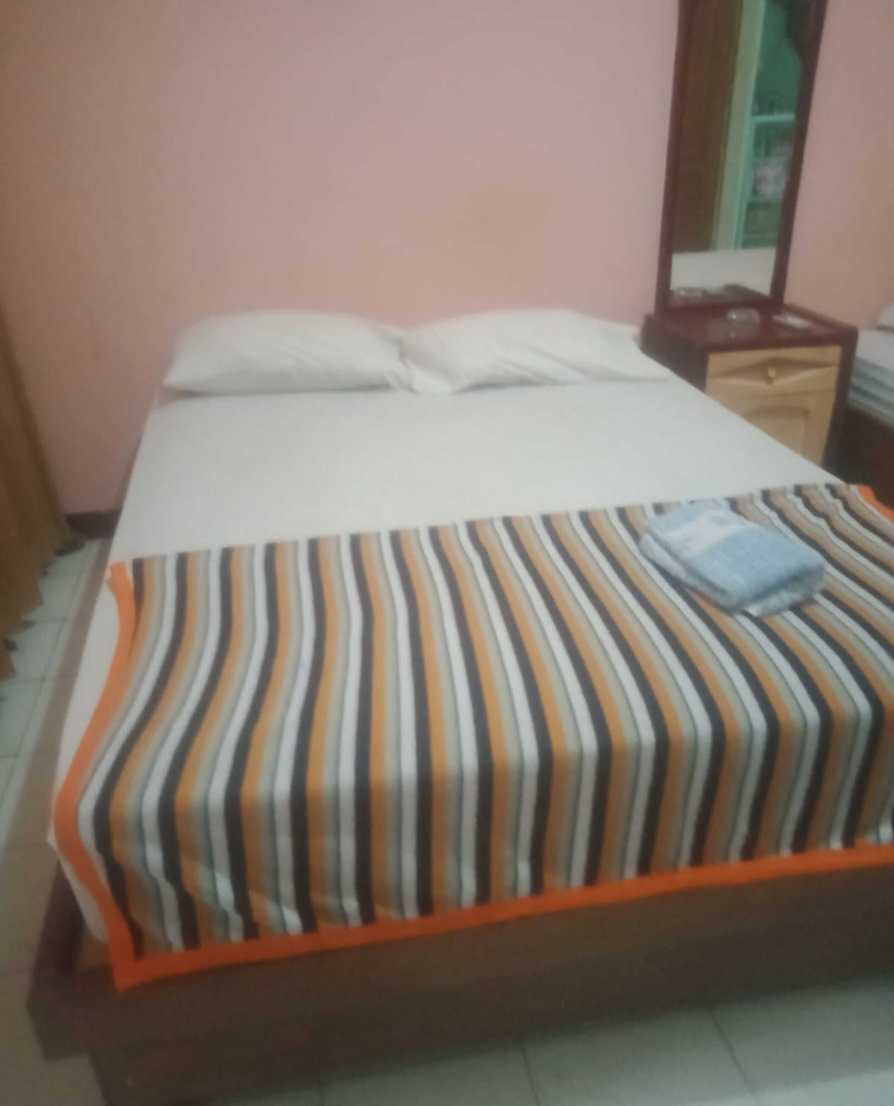 A bed with a striped bedspread and pillows, a wooden nightstand with a mirror, and a tiled floor.