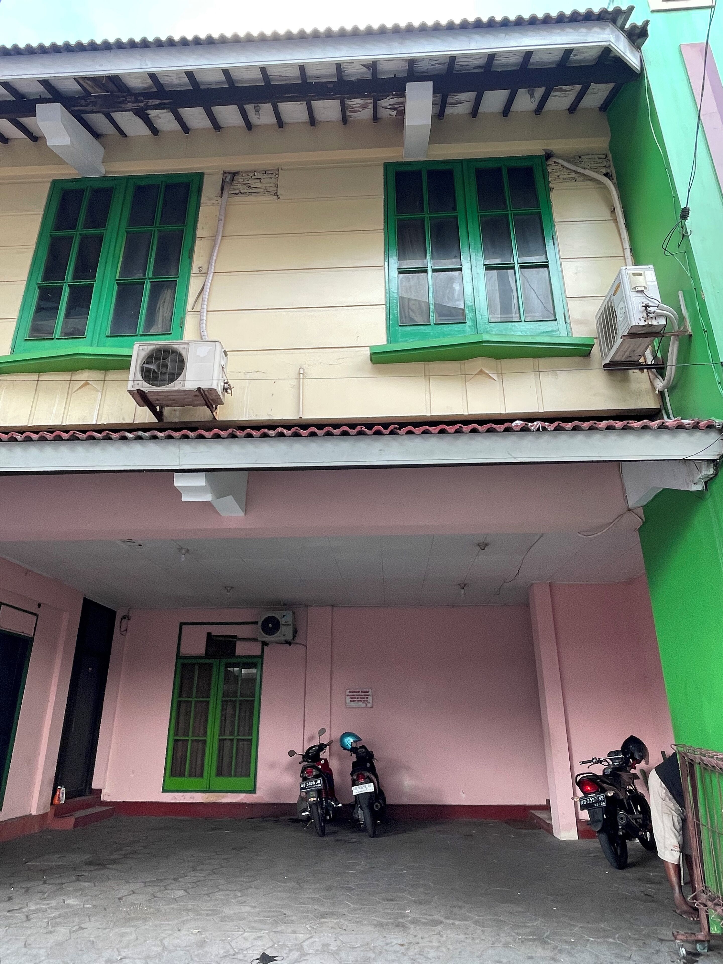 Un edificio con dos ventanas de marco verde, una unidad de aire acondicionado y un área de entrada cubierta donde se estacionan dos motocicletas.