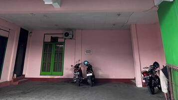Un edificio con dos ventanas de marco verde, una unidad de aire acondicionado y un área de entrada cubierta donde se estacionan dos motocicletas.