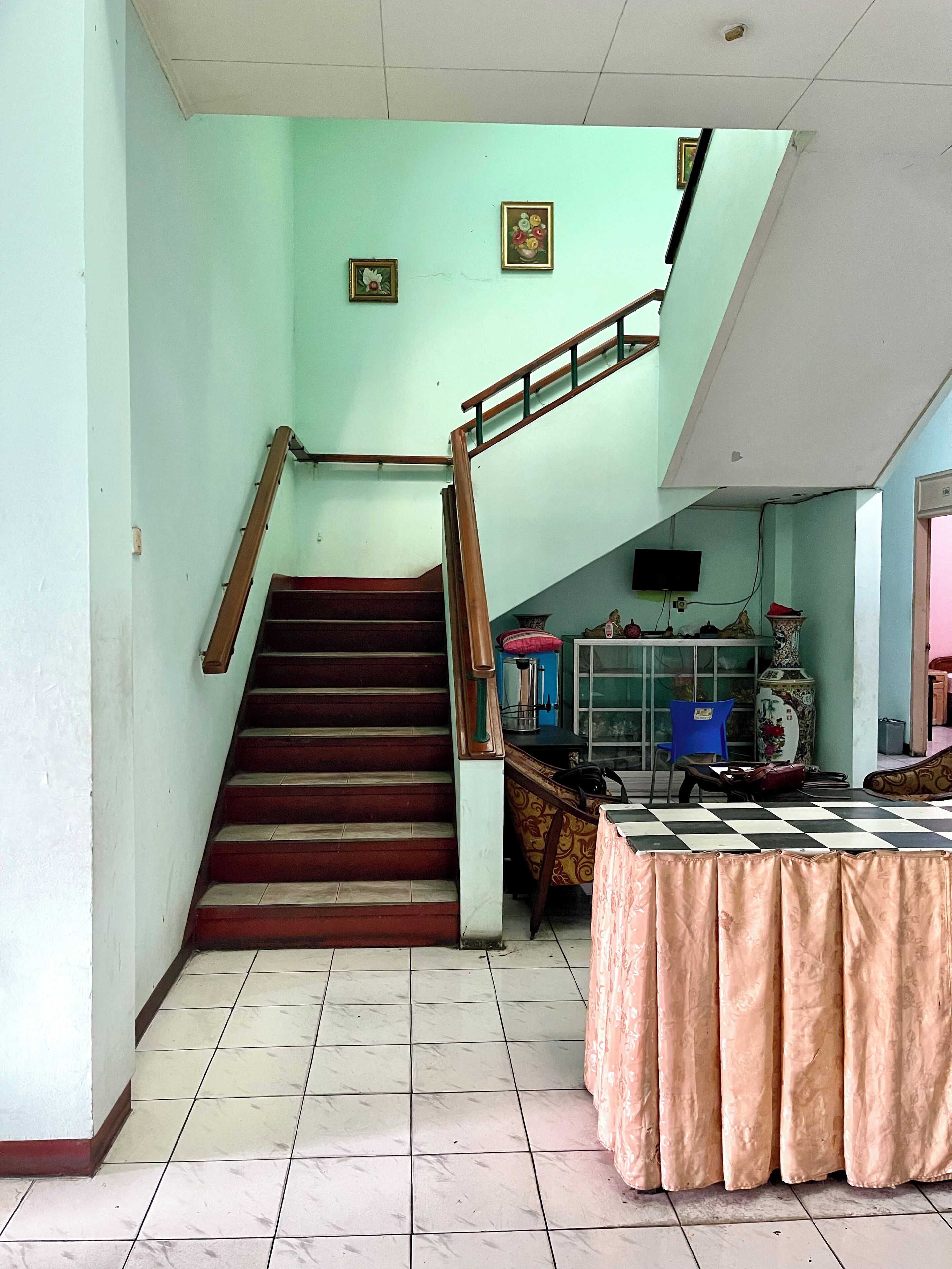 Una escalera que conduce a un piso superior, suelo de baldosas, una mesa con mantel rosa y un televisor en la pared.