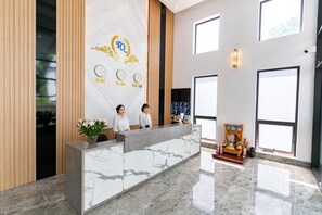 Uma área de recepção de hotel com balcão de mármore, duas recepcionistas, um relógio e uma parede decorativa com um logotipo.