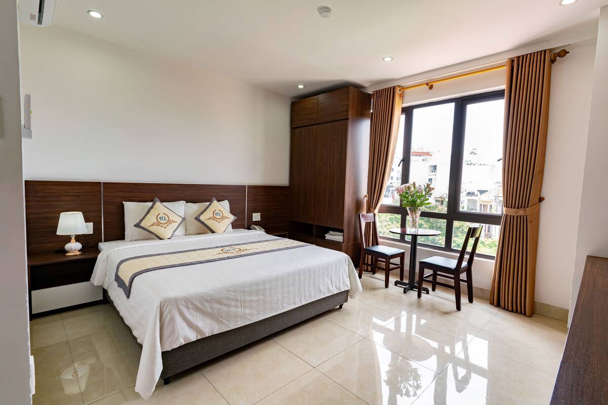 Une chambre d’hôtel avec un grand lit, une table de chevet avec une lampe, une armoire en bois, une fenêtre avec des rideaux et une petite table avec des chaises.