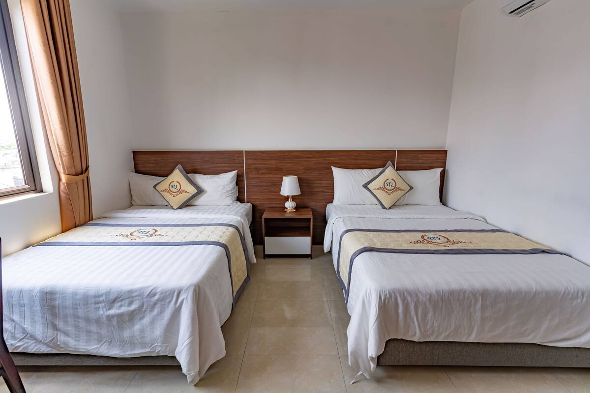 Une chambre d’hôtel avec deux lits, une table de chevet avec une lampe et une fenêtre avec des rideaux.