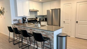 Una cocina moderna con gabinetes blancos, un refrigerador de acero inoxidable, una pared de ladrillo decorativa y taburetes de bar negros.