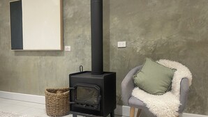 A room with a black wood-burning stove, a grey armchair with a green pillow, a wicker basket, and a framed picture on the wall.