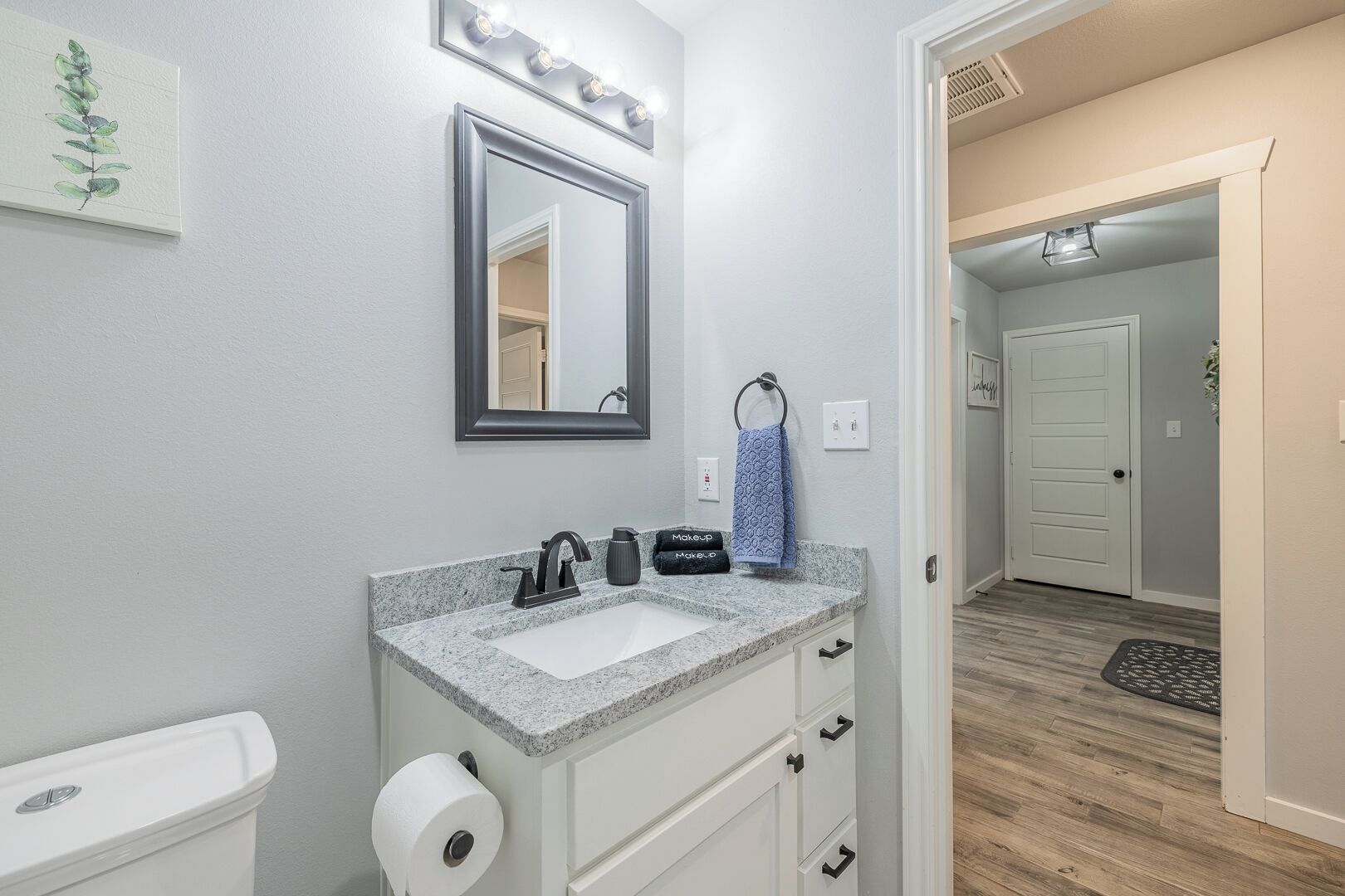 Une salle de bain avec un lavabo, un miroir et des toilettes. On y trouve un porte-serviettes avec une serviette bleue et une trousse de maquillage. Les murs sont peints en gris clair, et une porte donne accès à une autre pièce.