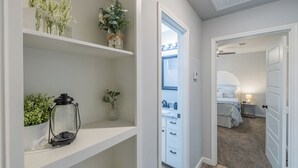 A hallway with white shelves holding plants and a lantern, leading to a bedroom with a bed, bedside table, and a window.