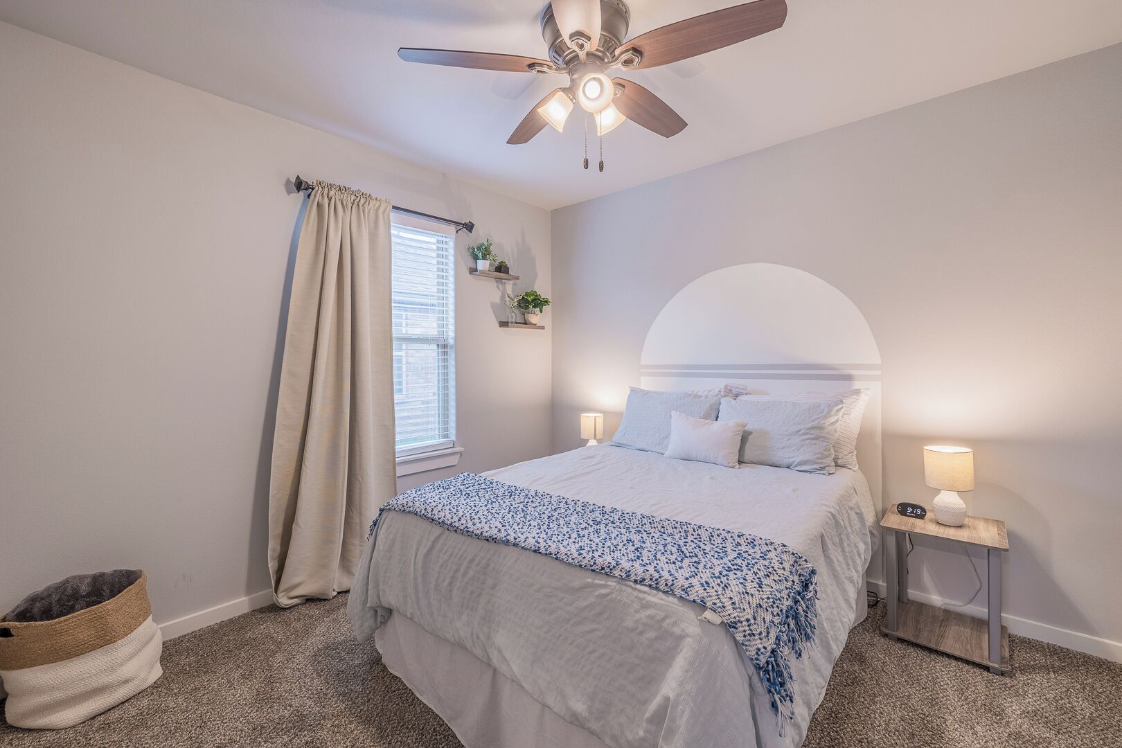 A bedroom with a bed, a ceiling fan, a bedside table with a lamp, and a window with curtains.
