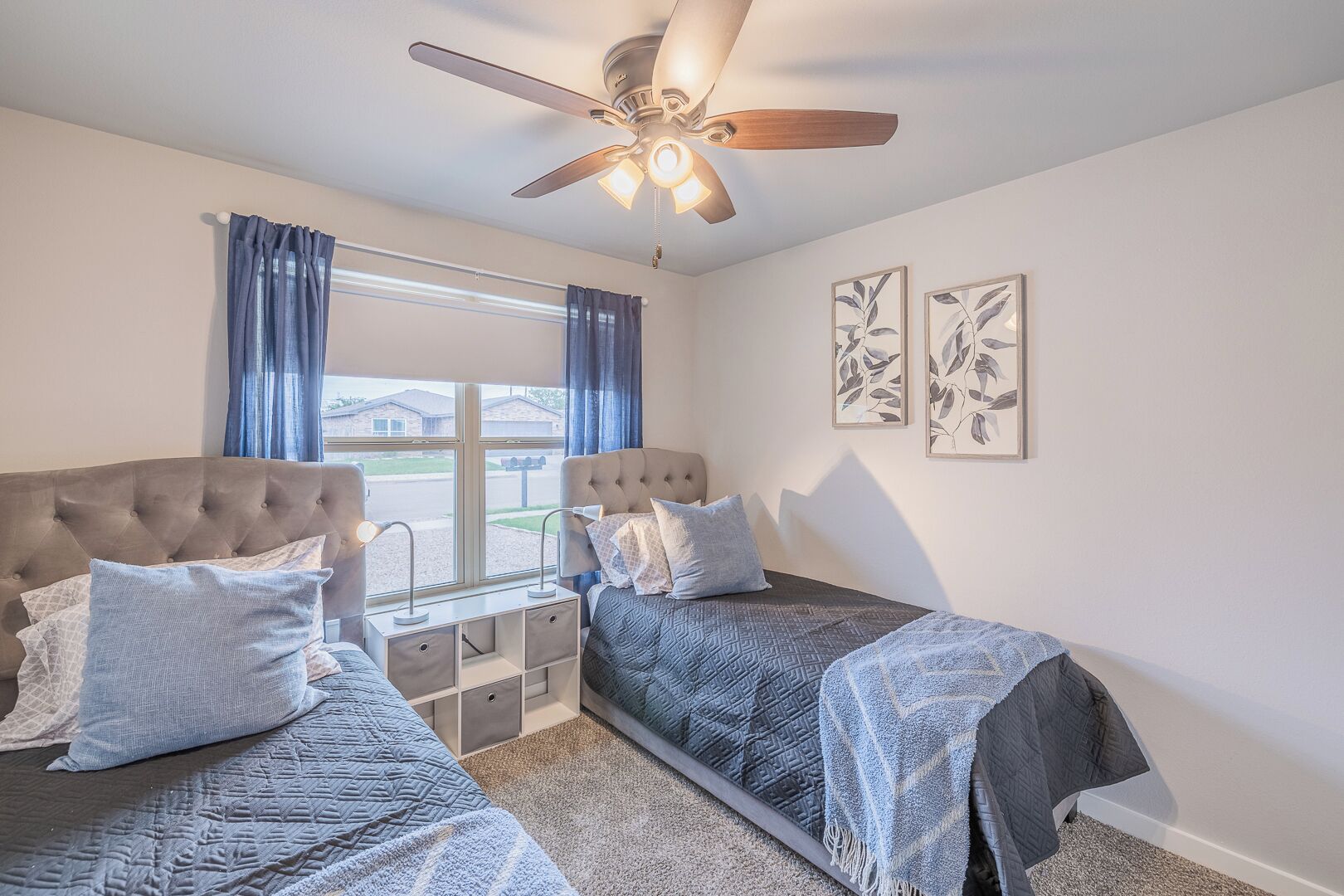 A bedroom with two beds, a ceiling fan, a window with curtains, and two framed leaf artworks on the wall.