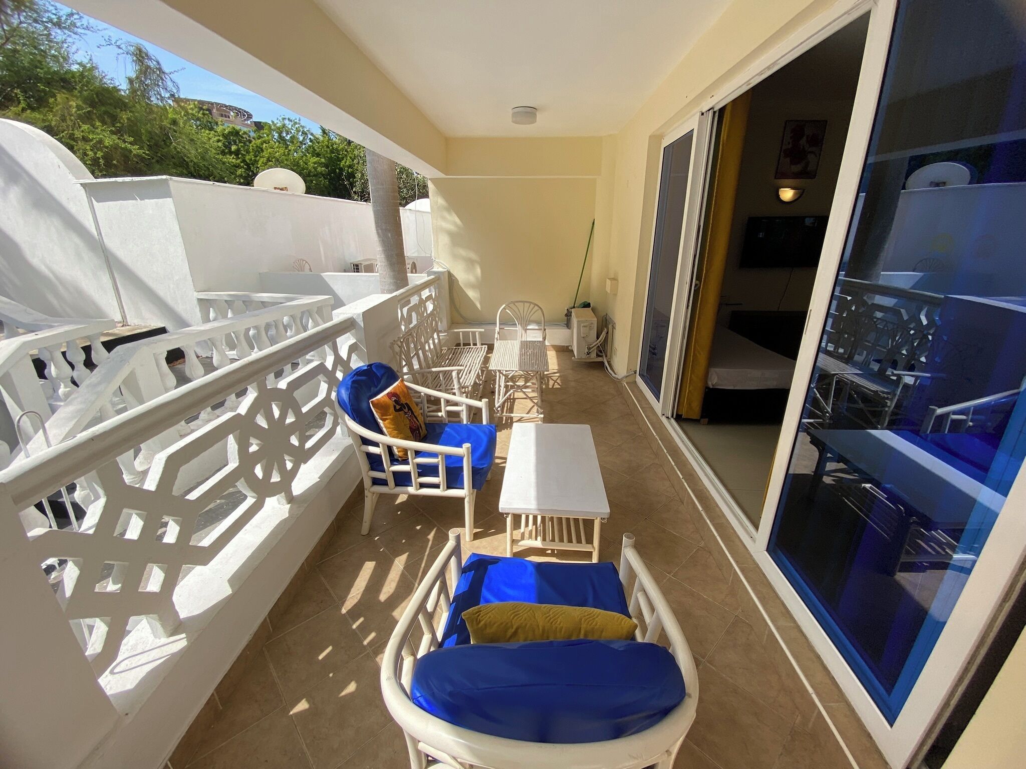 A balcony with white furniture, including chairs and a table, and a sliding glass door leading to a room with a bed and a chair.