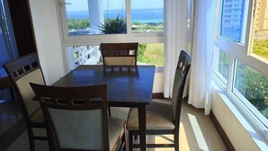 Un espace repas comprenant une table et des chaises, de grandes fenêtres offrant une vue sur la mer et la verdure, et un rideau blanc.