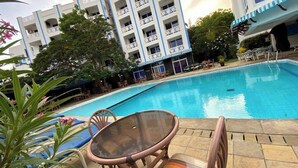 Une piscine entourée de chaises longues et d’une table, le tout bordé par un bâtiment doté de balcons et d’arbres.