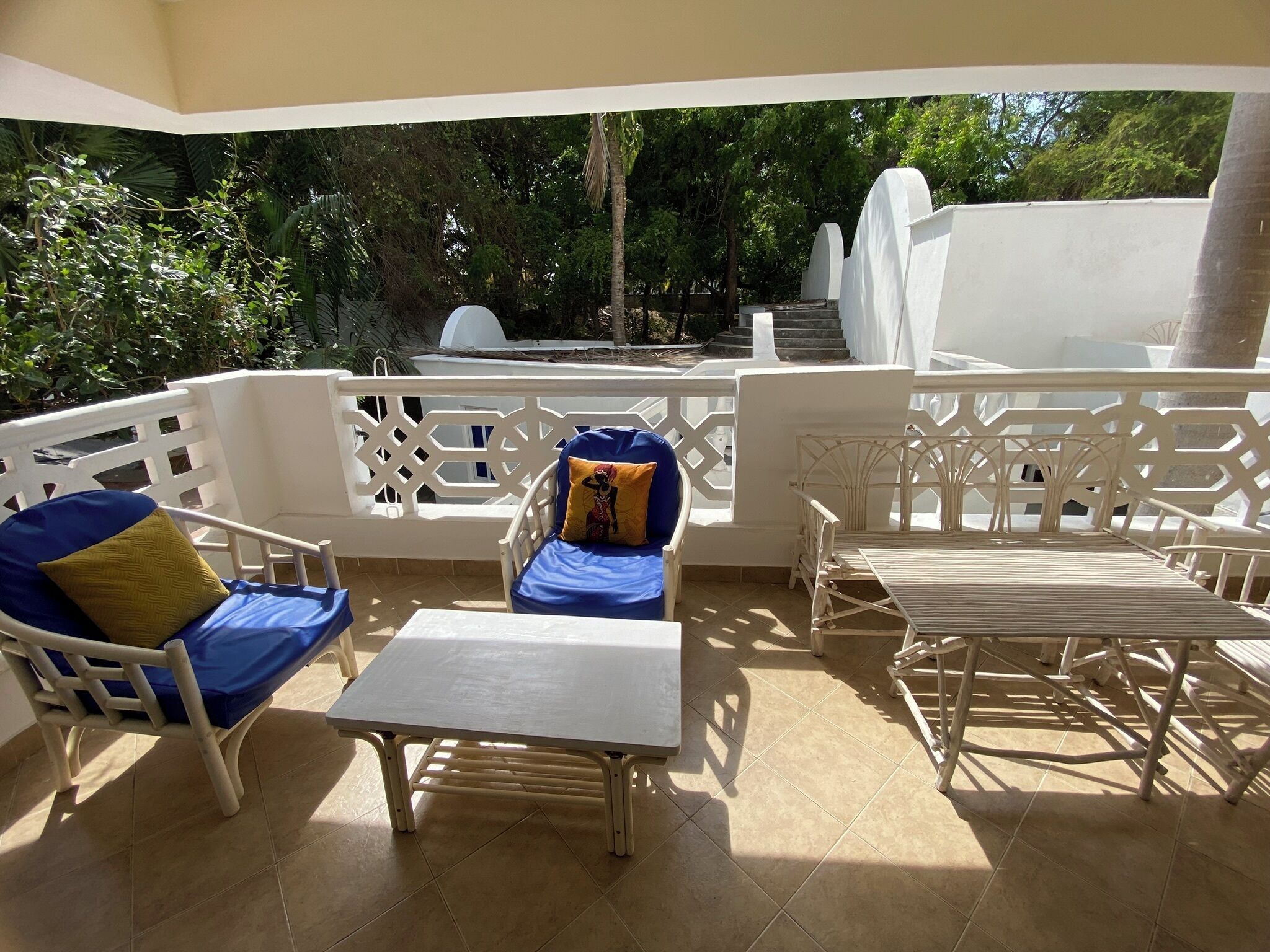 A covered outdoor seating area with white furniture, a small table, and a view of a pool and trees.