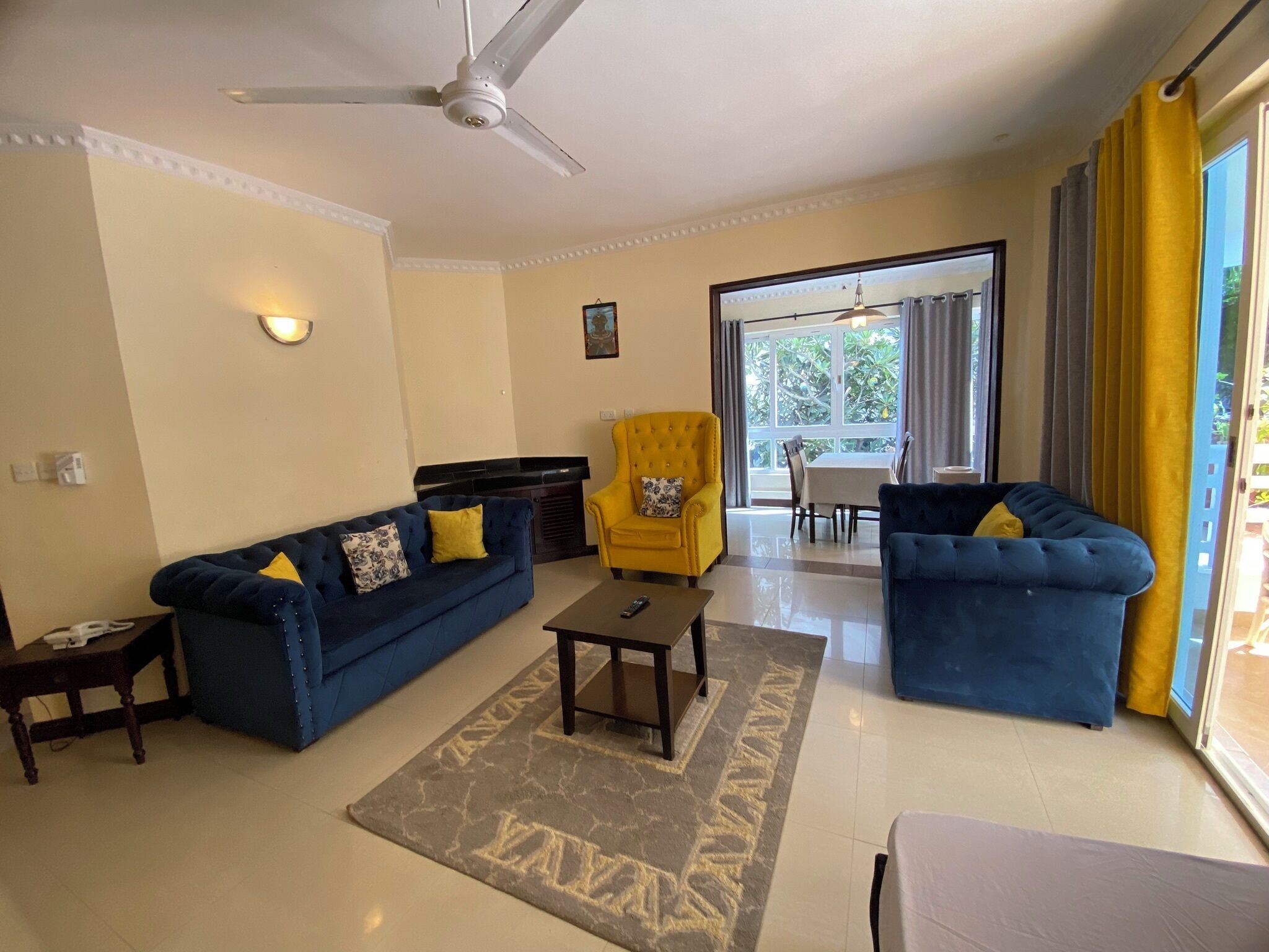 Una sala de estar con un sofá azul, un sillón amarillo y una mesa de centro. Hay un ventilador de techo, una ventana con cortinas y vistas al exterior.