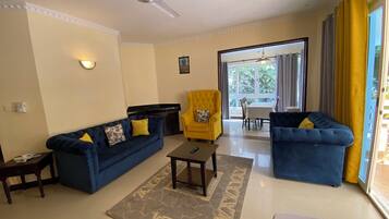 Una sala de estar con un sofá azul, un sillón amarillo y una mesa de centro. Hay un ventilador de techo, una ventana con cortinas y vistas al exterior.